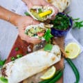 A hand holding a sandwich cut in half over a board of falafel sandwiches, lemon slices and green chillies