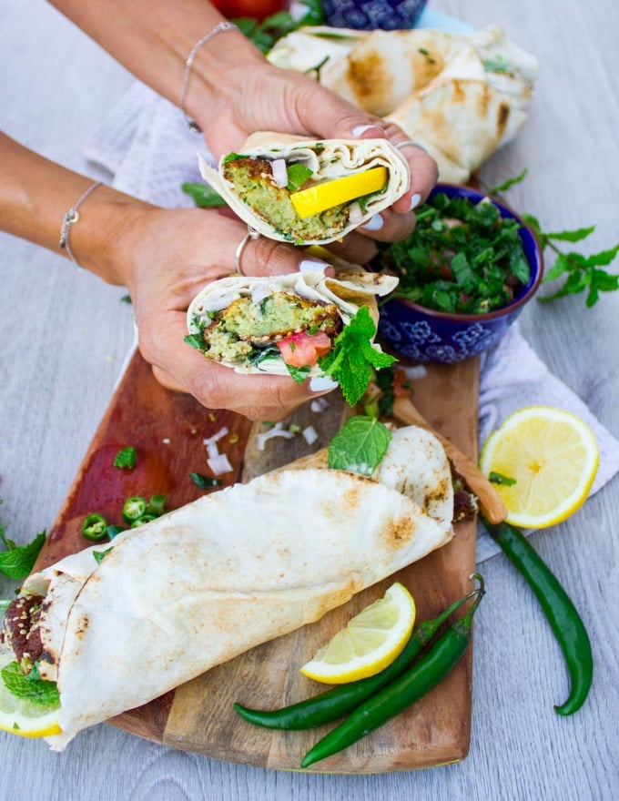 A hand holding a sandwich cut in half over a board of falafel sandwiches, lemon slices and green chillies
