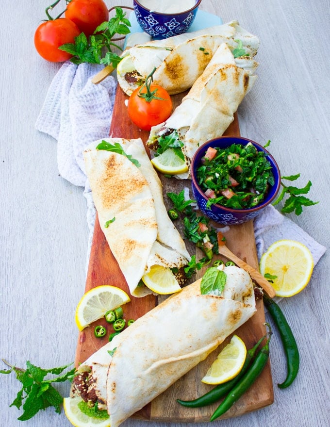 A set of four falafel sandwiches over a wooden board surrounded by lemon juice and tabouleh salad