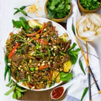 A table set up with a plate of stir fry recipe, chilli sauce on the same, cold drinks, extra cilantro and mint for garnish, chopsticks