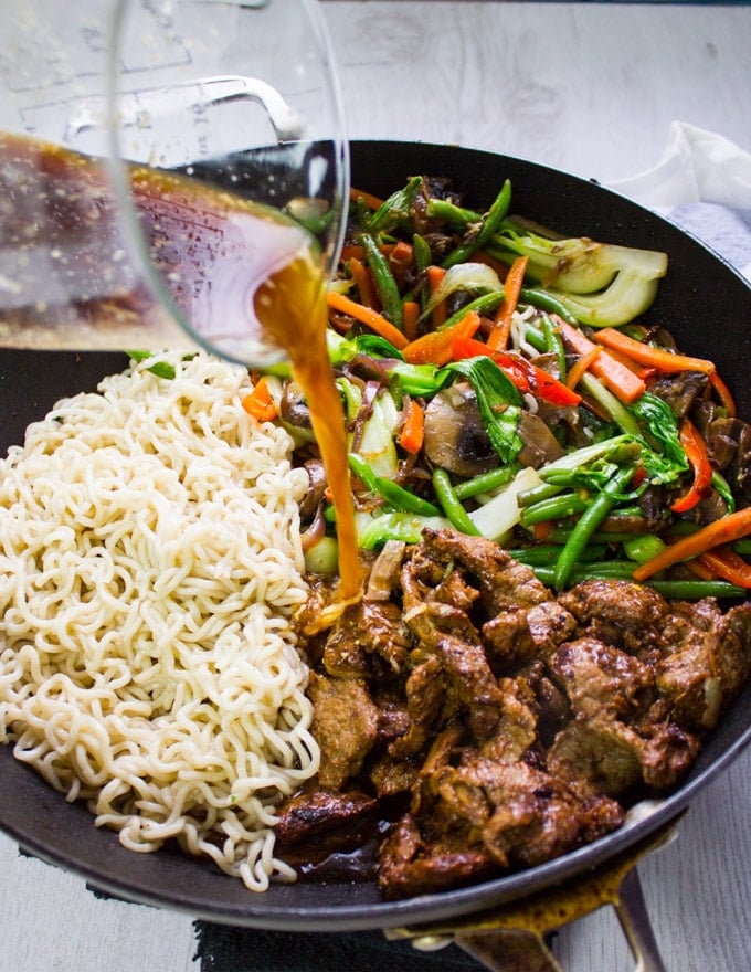 A wok with the ready stir fry veggies, lamb and noodles and the stir fry sauce being poured over them