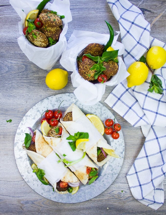 A plate of falafel wraps next to buckets of freshly fried falafel surrounded by mint, tomatoes and lemons