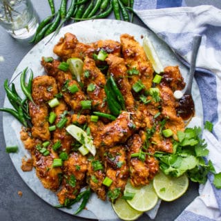 A plate of Korean Fried Chicken strips topped with cilantro and spread with lemon slices, green chillies and a kitchen towel.