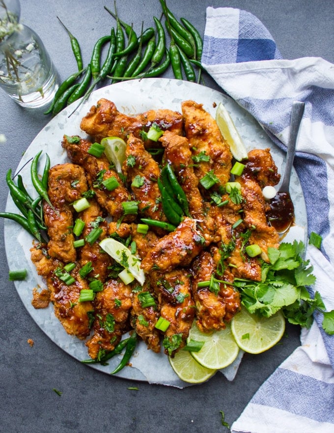 A plate of Korean Fried Chicken strips topped with cilantro and spread with lemon slices, green chillies and a kitchen towel.