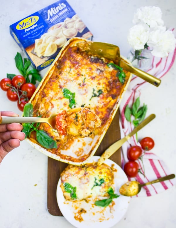 A hand holding a serving of homemade lasagna over a big dish of lasagna, surrounded by ripe tomatoes, a tea towel and flowers