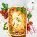 A big dish of homemade lasagna surrounded by a red tea towel, fresh basil leaves and a baby vine ripe tomatoes