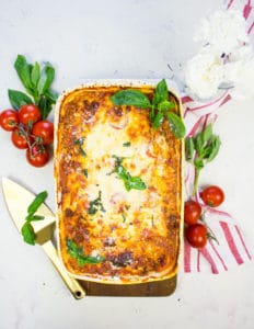 A big dish of homemade lasagna surrounded by a red tea towel, fresh basil leaves and a baby vine ripe tomatoes
