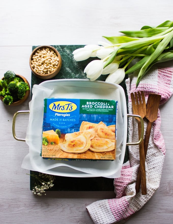 A box of frozen pierogies over a plate and surrounded by broccoli and pine nuts 