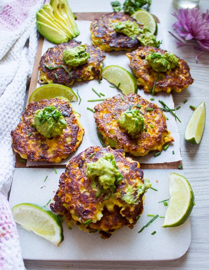 A white marle with lots of corn fritters spread out and topped with guacamole and separated by lime slices
