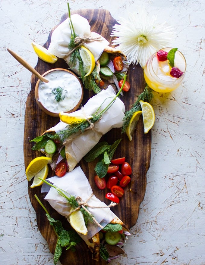 A platter of three gyros sandwiches with a bowl of tzatziki sauce and fresh tomatoes