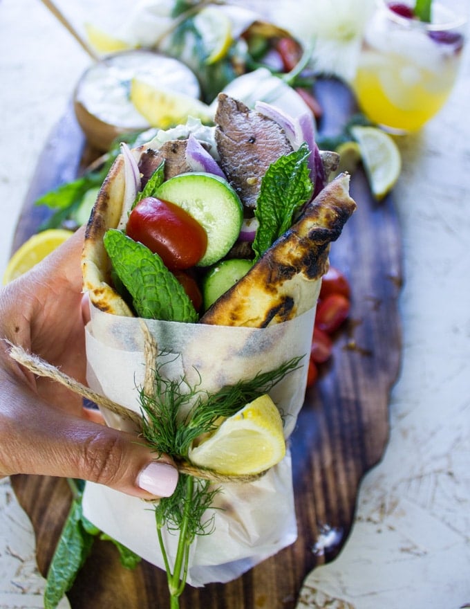 A hand holding a gyros sandwich wrapped and ready with lemon slices and showing the lamb, fresh veggies