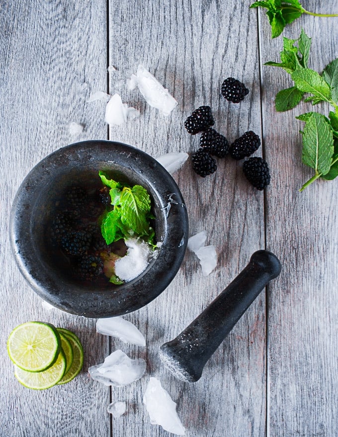 A mortar and pestle with blackberries, lime, sugar and mint surrounded by more mint, lime slices and ice 