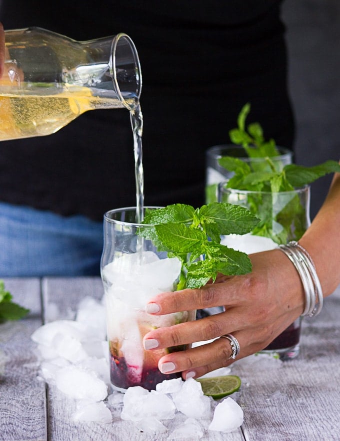 Pouring in the ginger ale/ lemon soda mixture while holding the mojitos cups with another hand to finish off the virgin mojitos recipe 