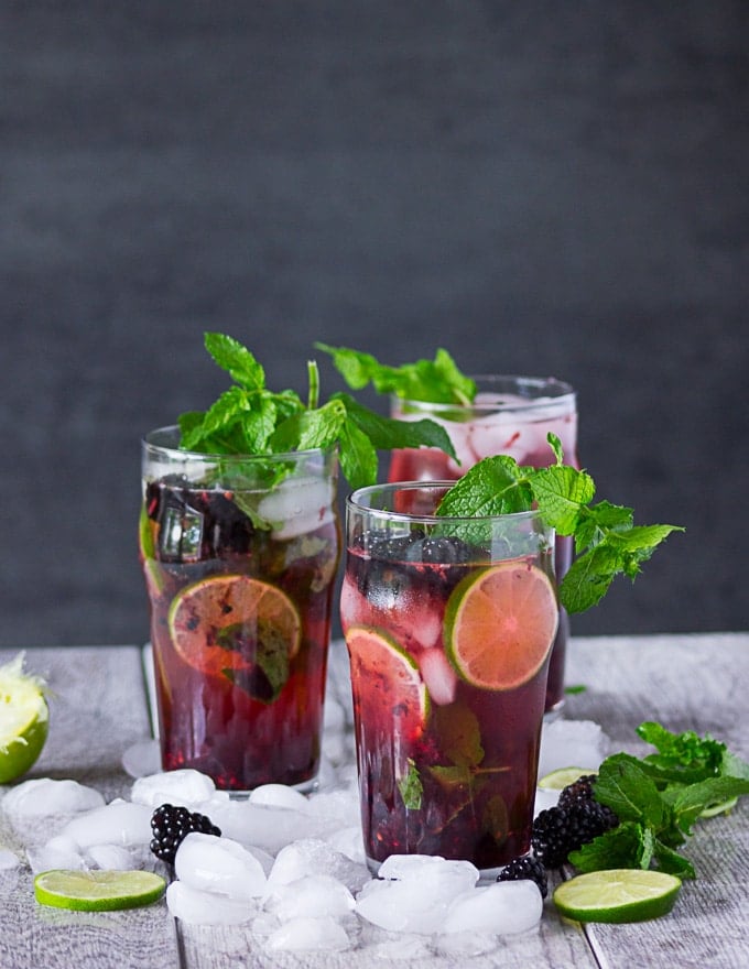 Three cups of prepared mojitos surrounded by ice and garnished with lime, mint and more blackberries