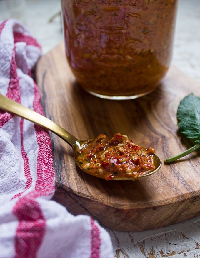 A spoon of hot sauce over a cutting board surrounded by basil and a tea towel 