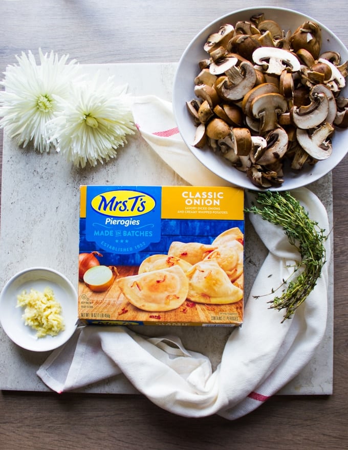 a box of Mrs.T's Pierogies - classic onion flavor surrounded by white flowers, a tea towel, a bowl of mushrooms and fresh thyme leaves