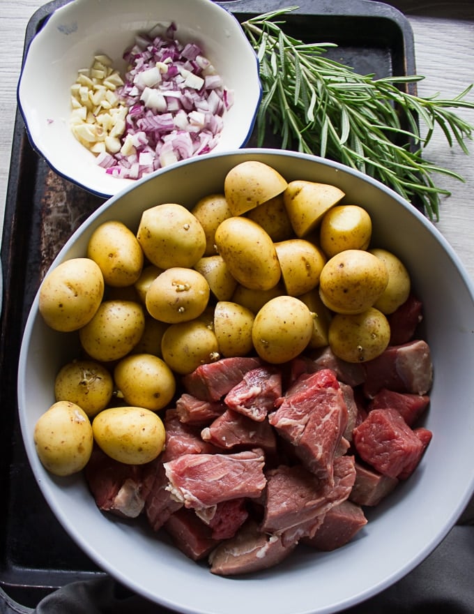 A bowl with potatoes cut up and lamb chunks cut up and another bowl with onions and garlic chopped up