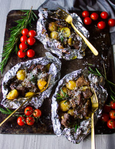 three packets of foil on the grill showing the lamb and potatoes with parmesan cheese, rosemary and tomatoes on the sides with three forks.