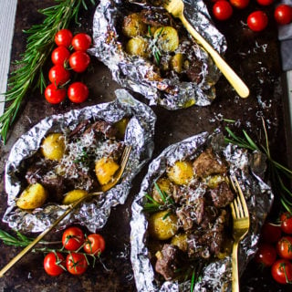 three packets of foil on the grill showing the lamb and potatoes with parmesan cheese, rosemary and tomatoes on the sides with three forks.