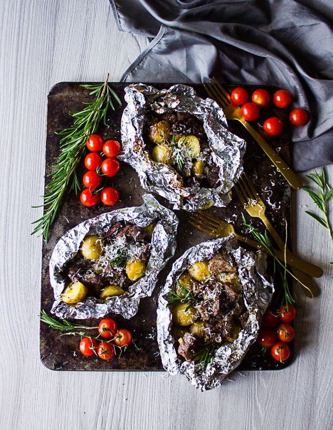 3 grill foil packets cooked and ready, opened up to expose the cooked lamb and potatoes with rosemary sprigs and baby tomatoes on the side.