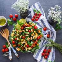 A salad with the halloumi cheese grilled and chopped, the tomatoes and arugula, the mint with lemon slices and the salad dressing on the side. Also a tea towel and flowers on the side.