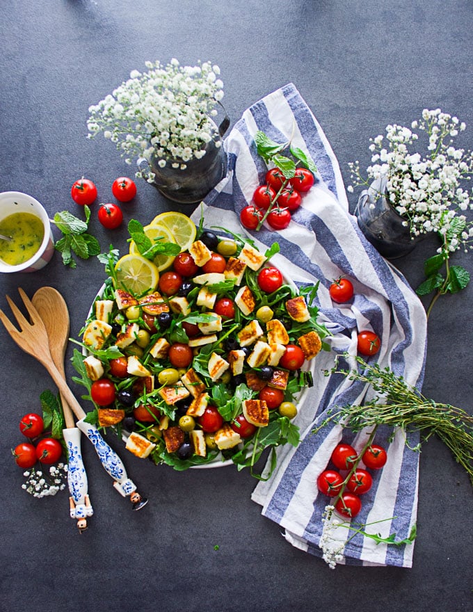 A salad with the halloumi cheese grilled and chopped, the tomatoes and arugula, the mint with lemon slices and the salad dressing on the side. Also a tea towel and flowers on the side.