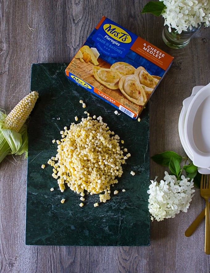 Ingredients for corn casseole over a marble board: the pierogies box, corn kernels, cream, ramekins and garlic