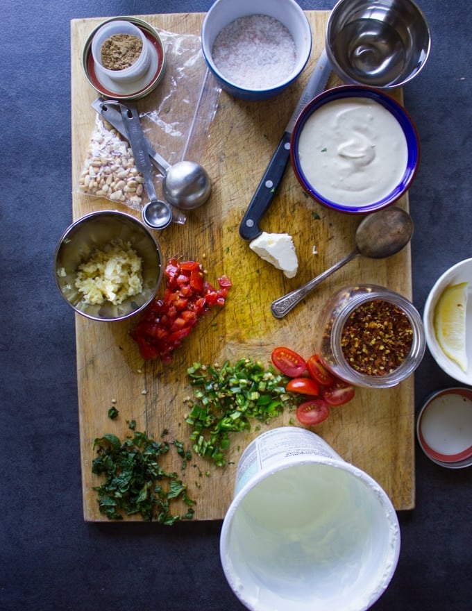 ingredients for eggplant salad on a cutting board, bowl of minced garlic, chopped tomatoes, bowl of vinegar, salt and spice, green chillies dced, pine nuts in a bowl , tahini sauce in a bowl, fresh chopped mint and parsley