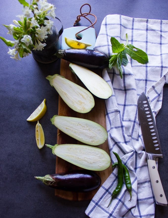 A sharp knife and eggplants sliced in half lengthwise on a cutting board surrounded by a tea towel