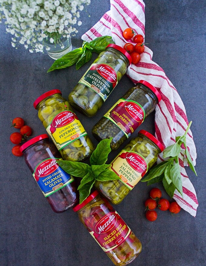 A collection of olives jars and capers surrounded by a tea towel