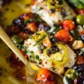 A spoon with the roast tomatoes and herbs and olives next to the baked fish
