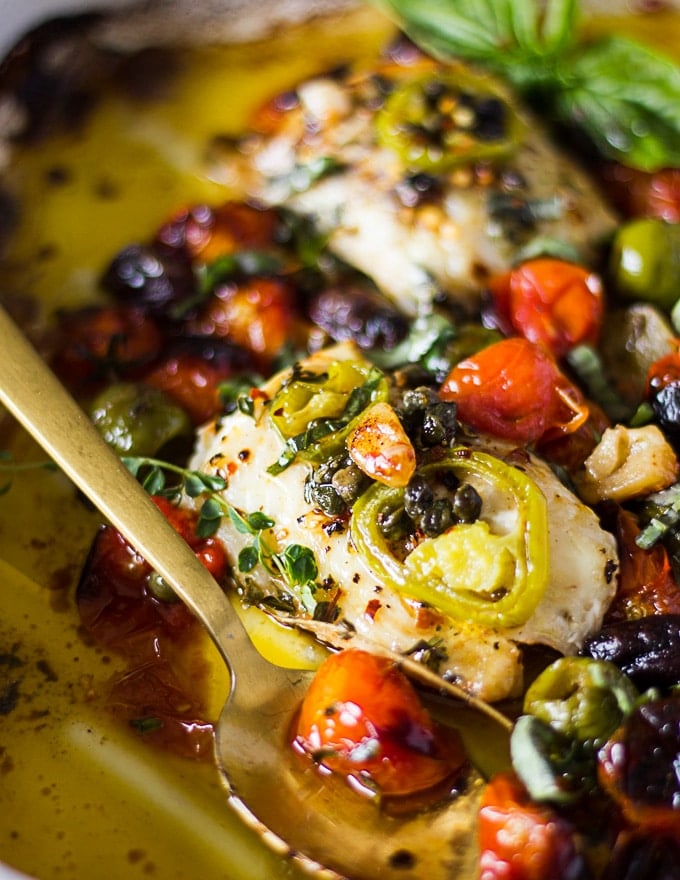 A spoon with the roast tomatoes and herbs and olives next to the baked fish