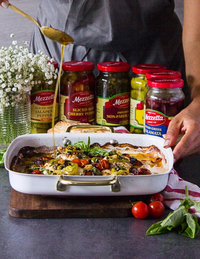 two hands holding the baked fish and a hand pouring the olive oil over the fish, the jars of olives and capers at the background