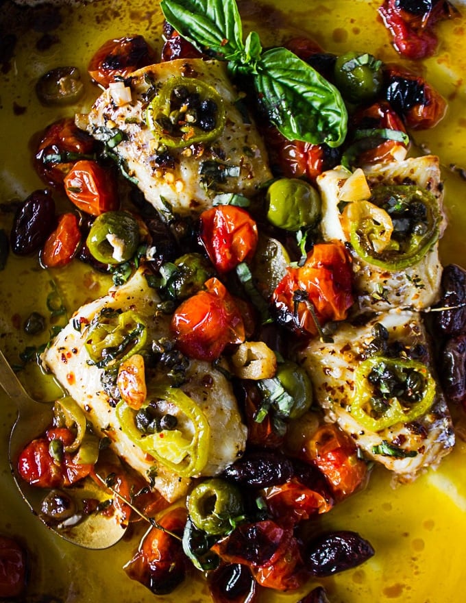 Close up of the final baked fish recipe out of the oven surrounded by olives, capers, roast tomatoes and herbs