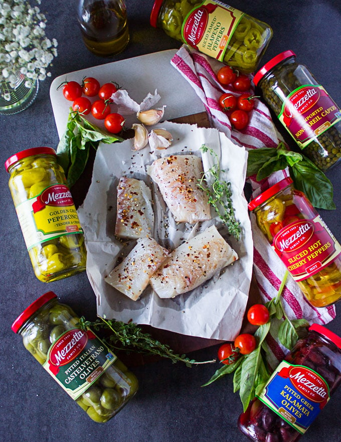 Haddock fillets seasoned on a pan and surrounded by garlic, fresh thyme, baby tomatoes and the jars of olives.