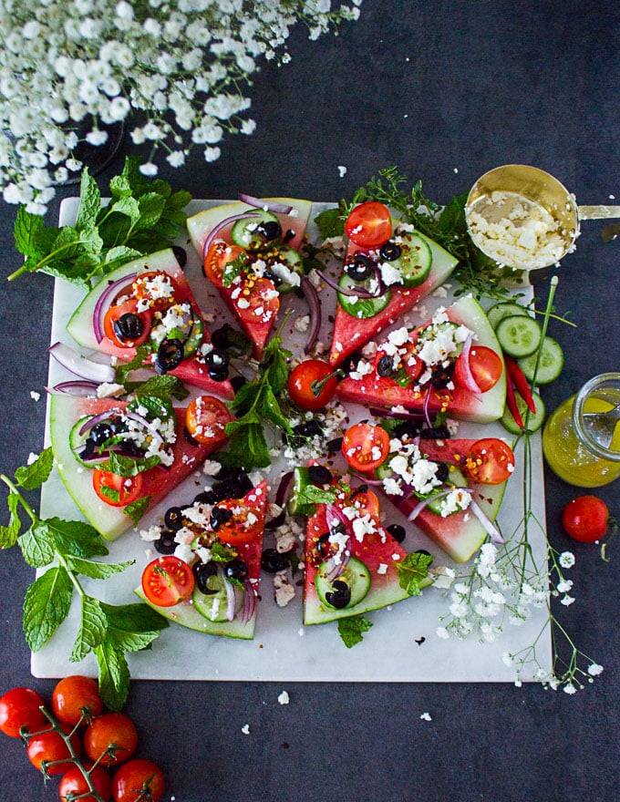 A white marble with watermelon salad loaded with Greek toppings surrounded by mint leaves and white flowers