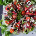 A whole disc of watermelon cut up into wedges and loaded with Greek salad toppings