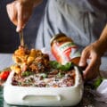 A hand serving the baked ziti right our of the pan using a spoon showing the cheesy baked ziti pasta and sauce