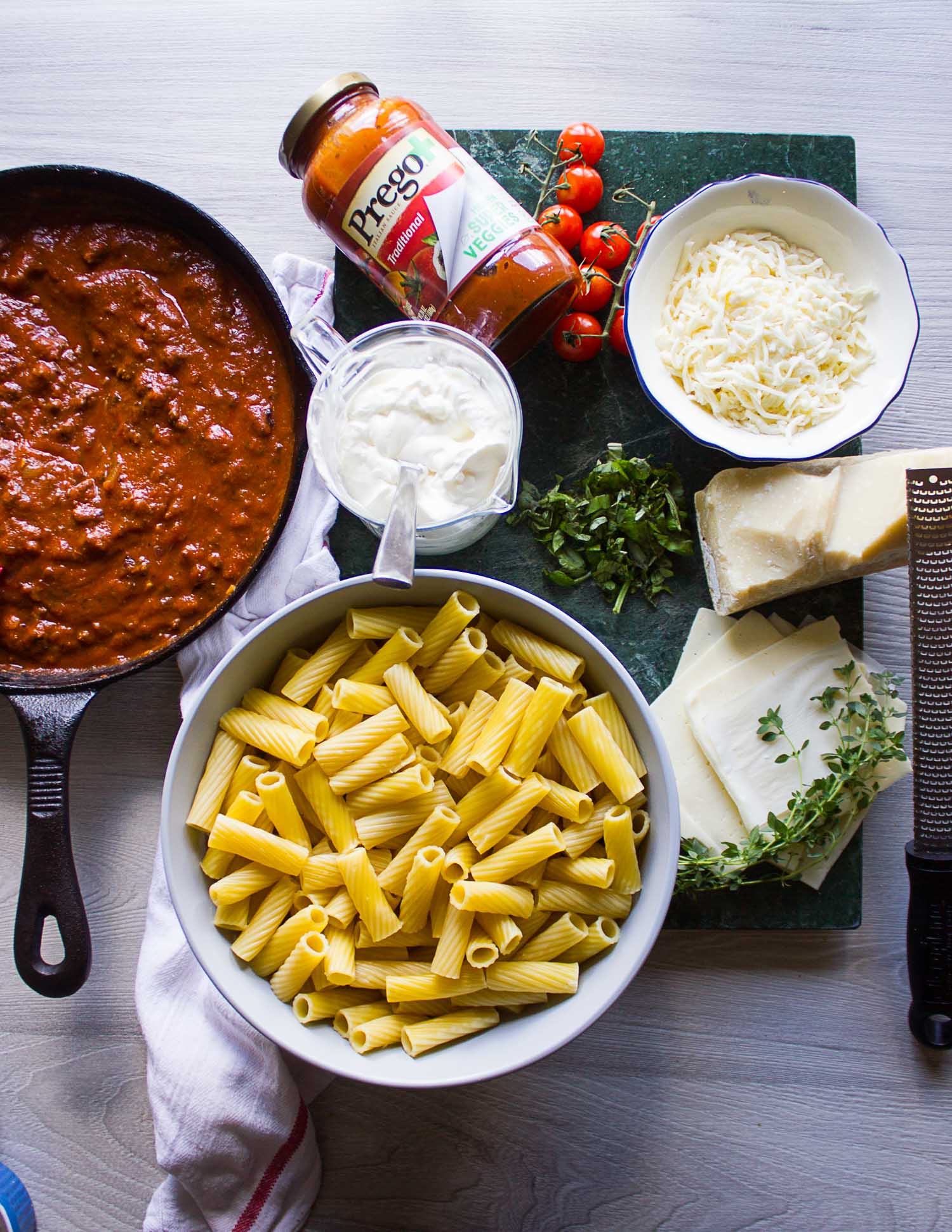 Ingredients for the baked ziti recipe on a board. Includes the cooked ziti pasta, the beef sauce, the cheeses and herbs