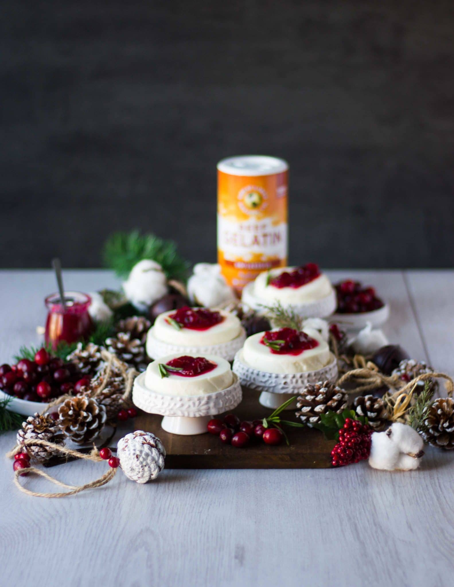 four individual serving panna cotta with cranberry sauce on a wooden board with a box of gelatin at the back and some fresh cranberries on the side