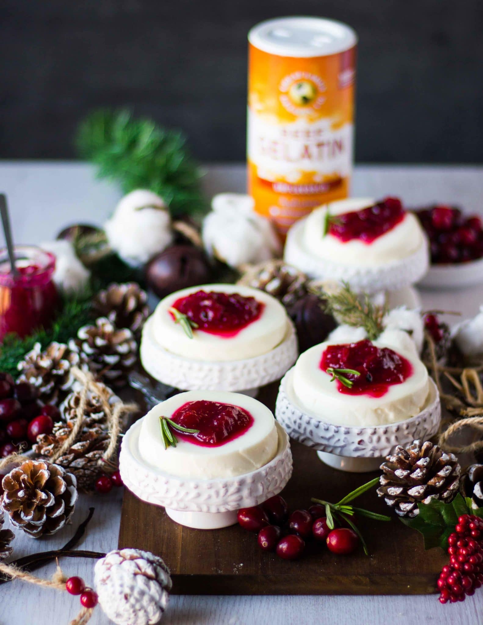 individual servings of four panna cotta recipe on serving plates finished with cranberry sauce and surrounded by fresh cranberries and wooden cones 