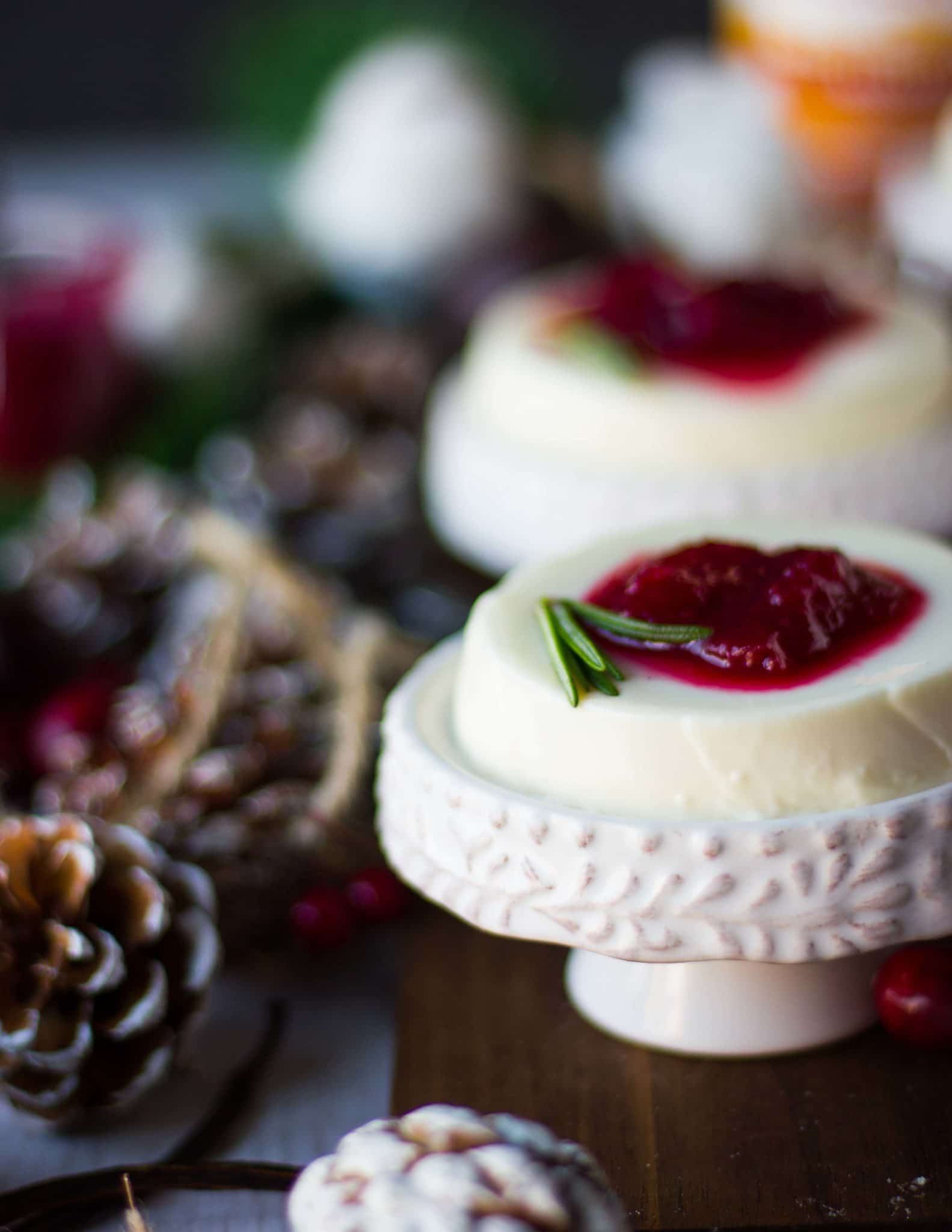 A single panna cotta on a serving plate over a wooden board next to a tree coe served with cranberry sauce and a sprig of rosemary