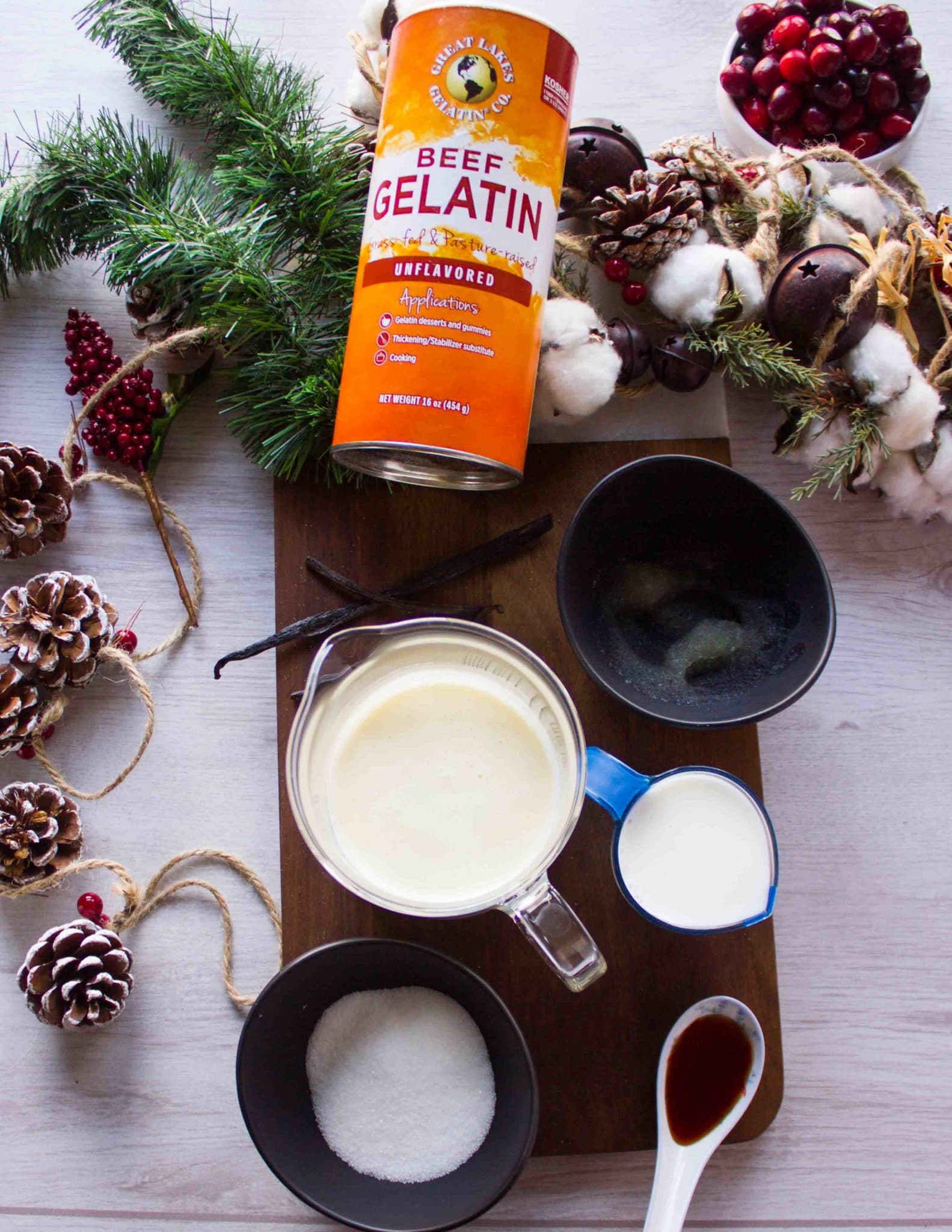 Ingredients for the panna cotta on a wodden board including cream in a pot, milk, sugar in a bowl, vanilla bean, vanilla extract, and gelatin in cold water 