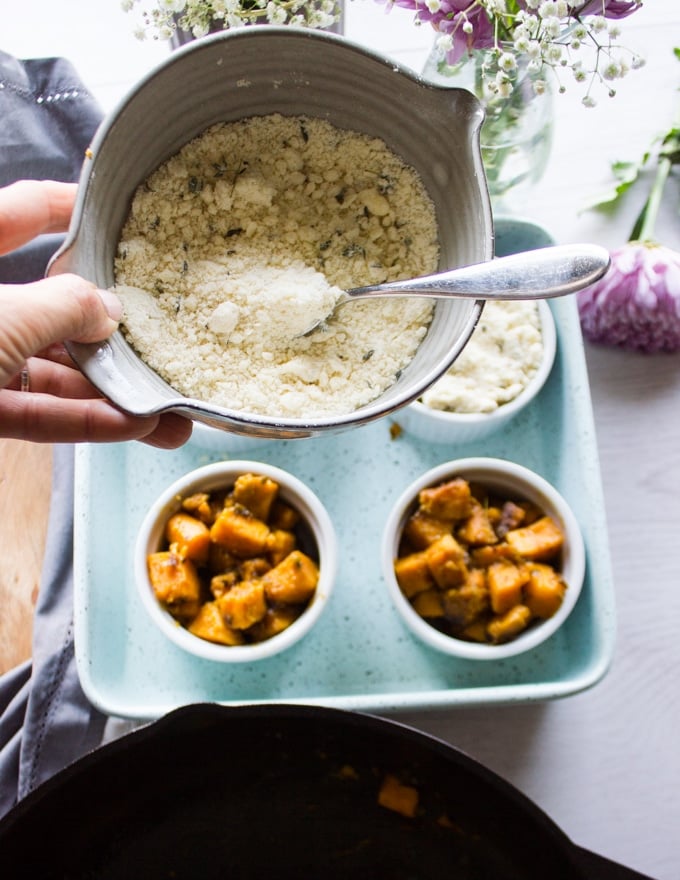 A hand holding the bowl of gratin topping over the ramekins with sweet potatoes ready to top with the crunchy topping