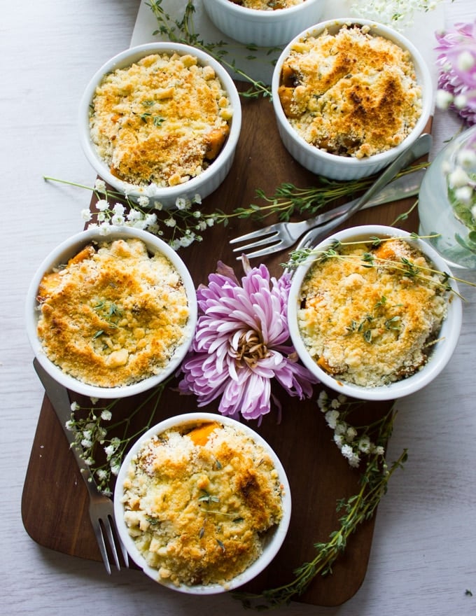 Top view of all the sweet potato gratins on a white table and a wooden cutting board surrounded by forks, fresh thyme leaves and a purple flower.