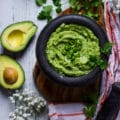 A bowl with guacamole recipe topped with chopped jalapenos and cilantro surrounded by hass avocados and an orange tea towel over a wooden board