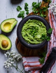 A bowl with guacamole recipe topped with chopped jalapenos and cilantro surrounded by hass avocados and an orange tea towel over a wooden board