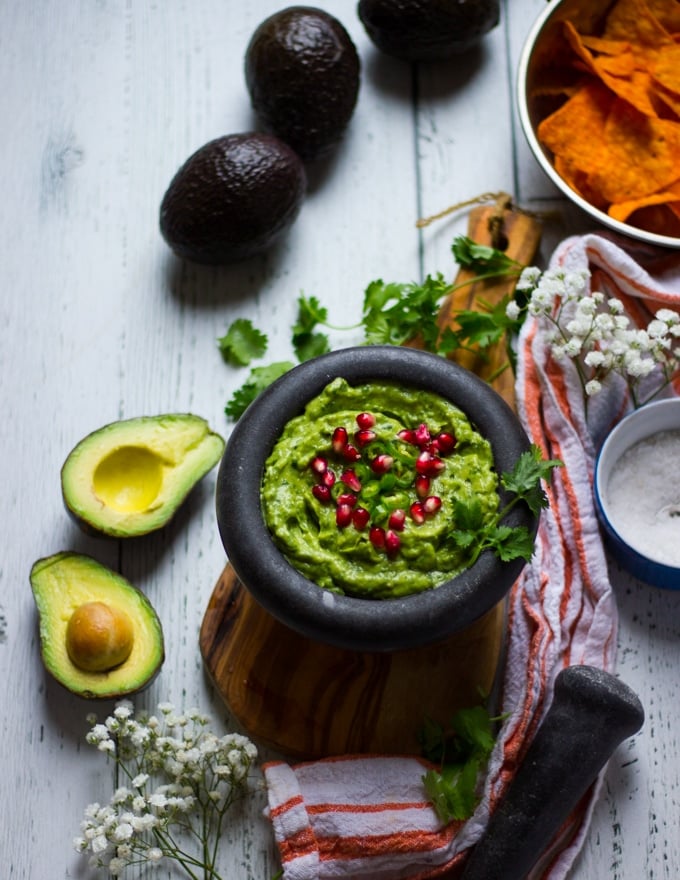 A guacamole recipe simple and topped with some jalapenos and pomegranate arils, not blended in.Some hass avocados around the bowl, a bowl of salt and a bowl of chips