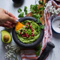 A hand holding a chip and dipping it into a guacamole dip in a bowl surrounded by a tea towel and hass avocados