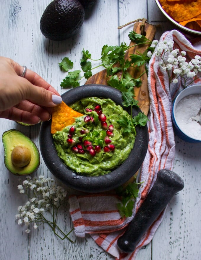 A hand holding a chip and dipping it into a guacamole dip in a bowl surrounded by a tea towel and hass avocados 